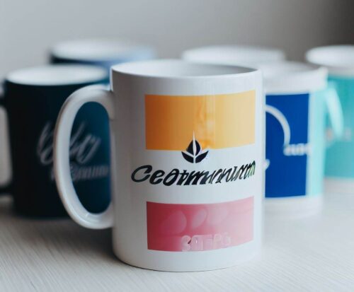 Bedruckte Keramiktasse mit farbigem Design und mehreren Tassen im Hintergrund auf hellem Tisch.