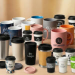 Eine Produktgruppenaufnahme auf einem Holztisch, die eine Vielzahl von gebrandeten Tassen und Mehrwegbechern zeigt. Dazu gehören Keramikbecher mit verschiedenen Farben und Mustern, Reisebecher aus Edelstahl, Keramik, Holzimitat und To-Go-Pappbecher mit individuellem Branding wie "Copenhagen 14" und "Ihr Logo".