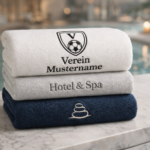 Drei hochwertige, gestapelte Handtücher mit individueller Bestickung für Verein, Hotel & Spa sowie Wellness auf einer Marmorablage vor einem Pool-Hintergrund.