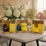 Jubiläums-Geschenkbox. Drei offene Kraftpapier-Geschenkboxen mit gebrandeten Werbemitteln von „DNZ NETWORKS“ (Flaschen, Notizbücher, Beutel, T-Shirt), platziert auf einem runden Bistrotisch vor einem großen Strauß Hortensien.