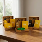 Drei offene Kraftpapier-Geschenkboxen mit gebrandeten Werbemitteln von „DNZ NETWORKS“, darunter Wasserflaschen (gelb, gold, schwarz), gelbe Notizbücher, ein T-Shirt, ein grünes Schlüsselband und ein Stift, auf einem Holztisch.